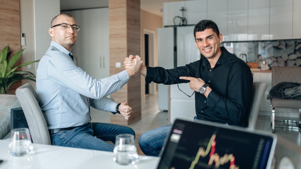 Dois homens de terno apertam as mãos em um ambiente de escritório moderno. Eles estão em uma conversa estratégica sobre investimentos, destacando a importância de tomar decisões financeiras bem-informadas. Ao fundo, uma tela de computador exibe gráficos financeiros complexos, com números em tempo real e indicadores de mercado, refletindo um cenário de trader ativo. O clima é de profissionalismo e colaboração, onde ambos estão alinhados em seus objetivos de crescimento financeiro, utilizando tecnologia de ponta para analisar e maximizar oportunidades de investimento. A cena transmite uma sensação de confiança e visão estratégica no mundo de investir.