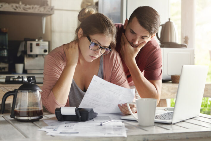 Casal jovem sentado à mesa da cozinha simbolizando inadimplência, olhando preocupado para contas e documentos, com uma calculadora, laptop e xícara de café à frente.